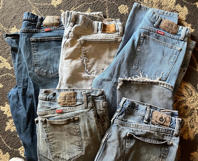以下が含まれることがあります： さまざまな青とグレーの色合いのデニムジーンズのコレクション。 バックポケット、ベルトループ、革パッチなどの詳細がいくつか見えます。 ダメージ加工が施されたものなど、使用感のあるジーンズもあります。