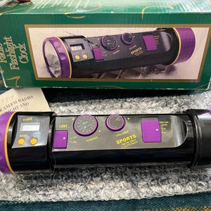 May include: Black and purple sports flashlight clock radio with a digital display, tuning and volume knobs. The box reads "Radio Flashlight Clock". The product is on bubble wrap.
