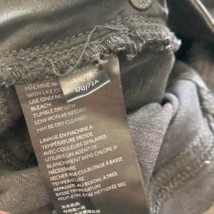 Pu&ograve; includere: Un'etichetta di cura nera con testo bianco che dettaglia le istruzioni di lavaggio. Il testo include "Machine Wash with Like Colors", "Use Only No Bleach", "Tumble Dry Low", "Low Iron as Needed", "May Be Dry Cleaned", "Lavage en Machine &agrave; Temp&eacute;rature Froide", "Avec Couleurs Similaires", "Utiliser un Agent de Blanchiment Sans Chlore Si N&eacute;cessaire", "S&eacute;cher par Culbutage &agrave; Basse Temp&eacute;rature", "Repasser, au Besoin, &agrave; Tr&egrave;s Basse Temp&eacute;rature", "Peut-&ecirc;tre &ecirc;tre Nettoy&eacute;e Sec", "冷水机洗", "与同色一起", "仅使用无需漂白剂", "低温烘干", "低温熨烫", "可干洗".