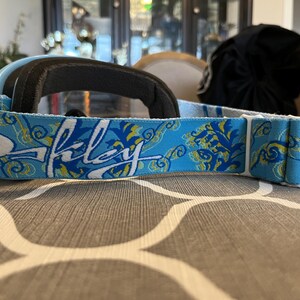 May include: Light blue Oakley ski goggles with a black padded frame and a patterned strap. The strap features the Oakley logo in white, along with a blue and yellow floral design. The goggles are designed for winter sports.