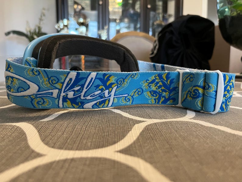 May include: Light blue Oakley ski goggles with a black padded frame and a patterned strap. The strap features the Oakley logo in white, along with a blue and yellow floral design. The goggles are designed for winter sports.