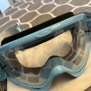 May include: Light blue Oakley ski goggles with a clear lens and a black foam padding. The strap is light blue with a white and yellow floral design. The goggles are designed for snow sports.