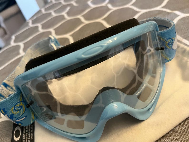 May include: Light blue Oakley ski goggles with a clear lens and a black foam padding. The strap is light blue with a white and yellow floral design. The goggles are designed for snow sports.