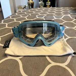 May include: Light blue ski goggles with clear lenses and a white and blue patterned strap. The goggles are resting on a white drawstring bag with a black tag. The goggles have a sporty design.