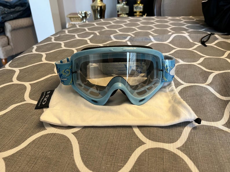 May include: Light blue ski goggles with clear lenses and a white and blue patterned strap. The goggles are resting on a white drawstring bag with a black tag. The goggles have a sporty design.