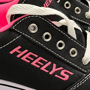 May include: Close-up of black Heelys roller skates with pink accents. The word "HEELYS" is embroidered in pink on the side of the shoe. White laces thread through silver eyelets. The sole is white with a black stripe.
