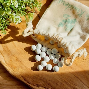 Könnte beinhalten: Ein weißer Baumwollbeutel mit Kordelzug, gefüllt mit kleinen weißen Keramikperlen. Der Beutel liegt auf einer Holzoberfläche.