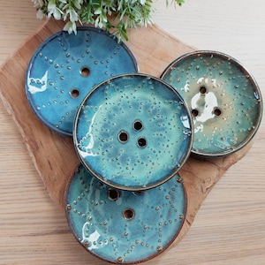 Op de afbeelding: Vier handgemaakte keramische zeepbakjes in tinten van blauwgroen en turquoise, elk met een uniek gespikkeld patroon en drie afvoergaten. De bakjes zijn gerangschikt op een houten plank, die hun glanzende afwerking en ambachtelijke kwaliteit benadrukt.