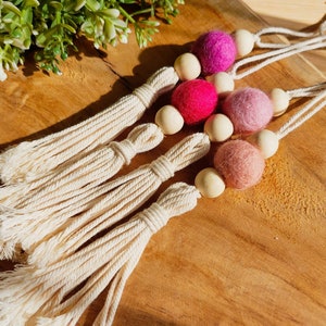 Op de afbeelding: Vier witte katoenen kwastjes met houten kralen en viltballen in tinten roze, rood en paars.