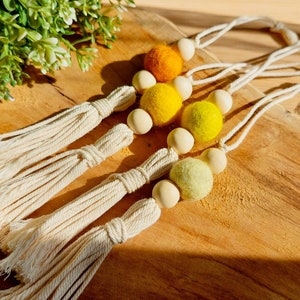 Op de afbeelding: Vier handgemaakte macramé wandhangers met houten kralen en viltballen in tinten van geel en oranje.