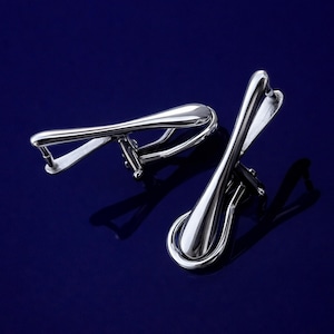 May include: Two silver metal objects, possibly jewellery or decorative items, are shown against a deep blue background. The objects have a curved, elongated design with a loop at the base and a smaller, angled element at the top. Shadows are cast by the lighting.