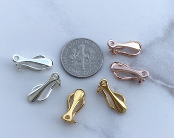 May include: A collection of six metal earring converters in silver, gold, and rose gold tones, arranged around a United States one dime coin for scale. Each converter features a hinged clip mechanism and a small loop for attaching jewelry.