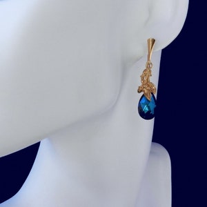 Pu&ograve; includere: Orecchino pendente color oro con una gemma blu scuro a forma di goccia. L'orecchino ha un design decorativo a foglia d'oro e un perno semplice. L'orecchino &egrave; esposto su un manichino bianco.