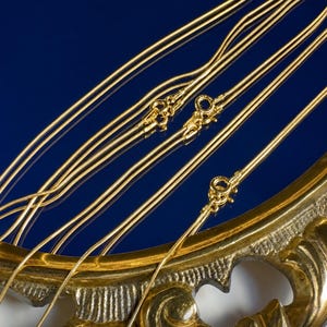 May include: Close-up of several gold-colored necklaces with delicate chains and ornate clasps. The necklaces are arranged against a deep blue background, with a decorative gold-colored frame in the foreground. The chains are thin and reflective.