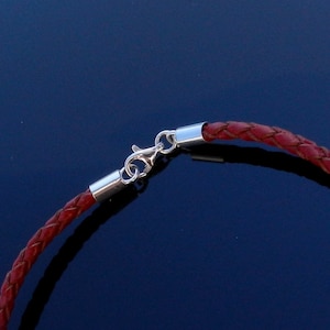Op de afbeelding: Close-up van een rood leren koordarmband met een zilveren sluiting. De armband is gevlochten en heeft een gepolijste zilveren sluiting.