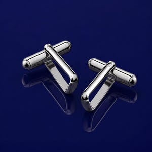 May include: A pair of shiny silver cufflinks with a curved, geometric design. The cufflinks have a polished finish and are set against a deep blue background, creating a sleek and modern aesthetic.