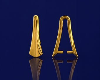Pode incluir: Dois brincos dourados de formato abstrato. Os brincos têm um acabamento liso e polido e uma superfície refletora. O design é geométrico, com um topo curvo e uma base alargada. O fundo é azul escuro.