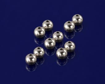 May include: A collection of shiny, silver-colored metal beads with a reflective surface. Each bead has a small hole. The beads are scattered on a dark blue background, creating a contrast that highlights their metallic sheen.