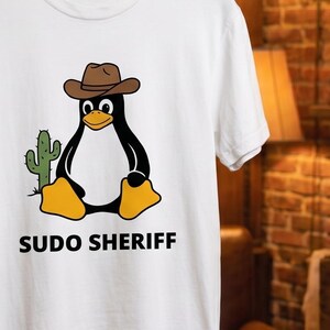 Puede incluir: Una camiseta blanca de manga corta con un gráfico de un pingüino blanco y negro que lleva un sombrero de vaquero marrón, sentado junto a un cactus verde. Debajo del pingüino, el texto SUDO SHERIFF está impreso en letras mayúsculas negras.