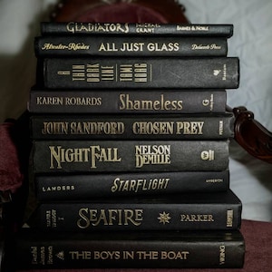 K&ouml;nnte beinhalten: Ein Stapel von zehn schwarzen Hardcover-B&uuml;chern mit goldenen Schriftz&uuml;gen auf den R&uuml;cken. Die Titel lauten "Gladiators", "All Just Glass", "Shameless", "Chosen Prey", "Nightfall", "Starflight", "Seafire" und "The Boys in the Boat".