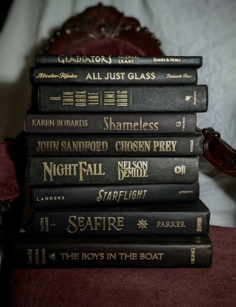 K&ouml;nnte beinhalten: Ein Stapel von zehn schwarzen Hardcover-B&uuml;chern mit goldenen Schriftz&uuml;gen auf den R&uuml;cken. Die Titel lauten "Gladiators", "All Just Glass", "Shameless", "Chosen Prey", "Nightfall", "Starflight", "Seafire" und "The Boys in the Boat".