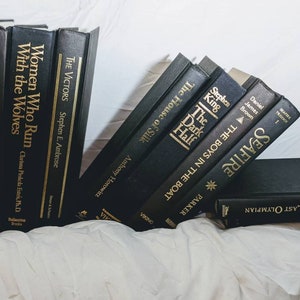 K&ouml;nnte beinhalten: Ein Stapel schwarzer gebundener B&uuml;cher mit goldenen Schriftz&uuml;gen auf den R&uuml;cken. Die Titel lauten "Die dunkle H&auml;lfte", "Die Jungs im Boot", "Seafire", "Der letzte Olympiasieger", "Die Sieger", "Frauen, die mit W&ouml;lfen rennen" und "Thesaurus".