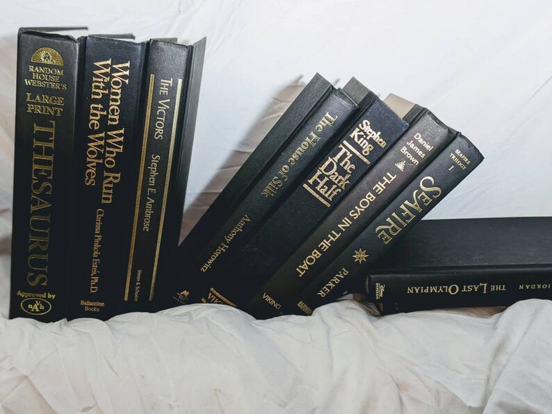 K&ouml;nnte beinhalten: Ein Stapel schwarzer gebundener B&uuml;cher mit goldenen Schriftz&uuml;gen auf den R&uuml;cken. Die Titel lauten "Die dunkle H&auml;lfte", "Die Jungs im Boot", "Seafire", "Der letzte Olympiasieger", "Die Sieger", "Frauen, die mit W&ouml;lfen rennen" und "Thesaurus".