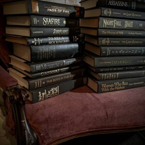 K&ouml;nnte beinhalten: Ein Stapel gebundener B&uuml;cher mit schwarzen Einb&auml;nden und goldenen Schriftz&uuml;gen. Die B&uuml;cher sind in einem B&uuml;cherregal angeordnet, wobei die Titel nach au&szlig;en zeigen. Zu den Titeln geh&ouml;ren "Seafire", "To Kill a Mockingbird", "The Boys in the Boat" und "Women Who Run With the Wolves".