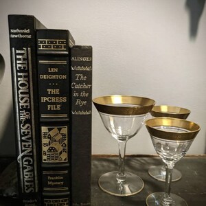 K&ouml;nnte beinhalten: Drei schwarze Hardcover-B&uuml;cher mit goldenen Schriftz&uuml;gen auf einer dunklen Oberfl&auml;che. Die B&uuml;cher sind betitelt mit "The House of the Seven Gables", "The Ipcress File" und "The Catcher in the Rye". Vor den B&uuml;chern stehen drei goldger&auml;nderte Stielgl&auml;ser.