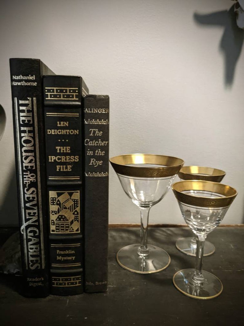 K&ouml;nnte beinhalten: Drei schwarze Hardcover-B&uuml;cher mit goldenen Schriftz&uuml;gen auf einer dunklen Oberfl&auml;che. Die B&uuml;cher sind betitelt mit "The House of the Seven Gables", "The Ipcress File" und "The Catcher in the Rye". Vor den B&uuml;chern stehen drei goldger&auml;nderte Stielgl&auml;ser.