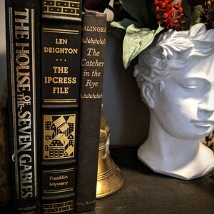 K&ouml;nnte beinhalten: Drei gebundene B&uuml;cher mit goldenen Schriftz&uuml;gen auf schwarzen Buchr&uuml;cken. Die Titel lauten "The House of Seven Gables" von Nathaniel Hawthorne, "The Ipcress File" von Len Deighton und "The Catcher in the Rye" von J.D. Salinger. Die B&uuml;cher sind vertikal auf einer dunklen Oberfl&auml;che angeordnet.