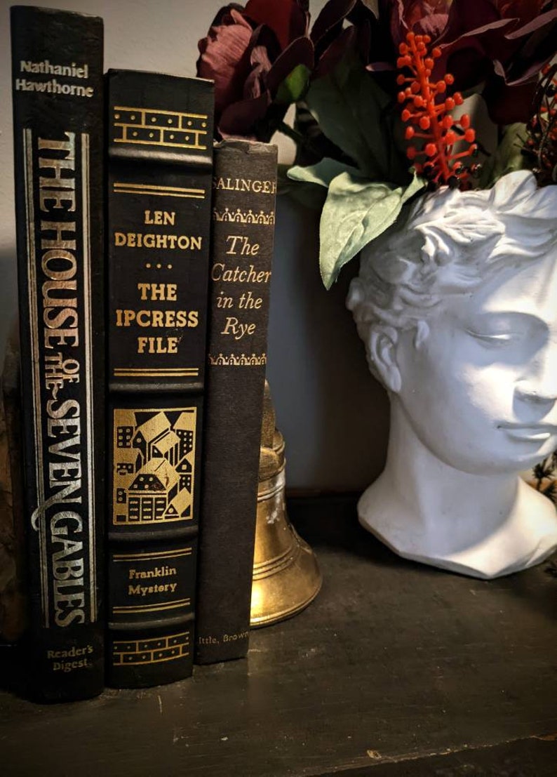 K&ouml;nnte beinhalten: Drei gebundene B&uuml;cher mit goldenen Schriftz&uuml;gen auf schwarzen Buchr&uuml;cken. Die Titel lauten "The House of Seven Gables" von Nathaniel Hawthorne, "The Ipcress File" von Len Deighton und "The Catcher in the Rye" von J.D. Salinger. Die B&uuml;cher sind vertikal auf einer dunklen Oberfl&auml;che angeordnet.