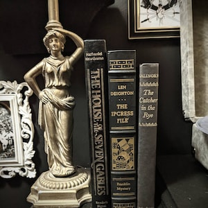 K&ouml;nnte beinhalten: Drei gebundene B&uuml;cher auf einer dunklen Oberfl&auml;che. Die B&uuml;cher tragen die Titel "The House of the Seven Gables" von Nathaniel Hawthorne, "The Ipcress File" von Len Deighton und "The Catcher in the Rye" von J.D. Salinger. Die B&uuml;cher sind alle schwarz mit goldenen Schriftz&uuml;gen und Illustrationen.