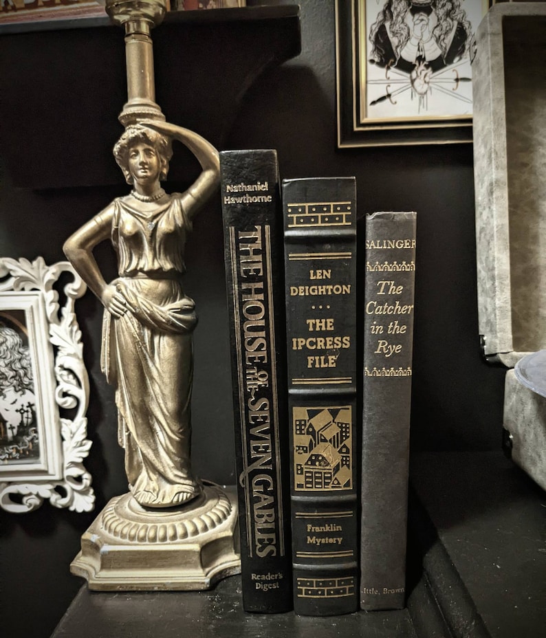 K&ouml;nnte beinhalten: Drei gebundene B&uuml;cher auf einer dunklen Oberfl&auml;che. Die B&uuml;cher tragen die Titel "The House of the Seven Gables" von Nathaniel Hawthorne, "The Ipcress File" von Len Deighton und "The Catcher in the Rye" von J.D. Salinger. Die B&uuml;cher sind alle schwarz mit goldenen Schriftz&uuml;gen und Illustrationen.