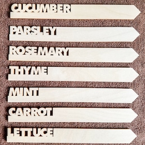May include: Seven wooden garden markers with cut-out lettering. The markers are labeled with the names of herbs and vegetables: Cucumber, Parsley, Rosemary, Thyme, Mint, Carrot, and Lettuce. The markers are light brown and have an arrow shape.