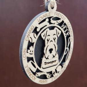 May include: A wooden ornament with a silhouette of a dog's head and the words "Loyal, Caring, Lovable, Friendly, Warm, Adorable, Dependable, Joyous, Companion, Woof" around the edge. The dog's name "Trooper" is written on a tag.
