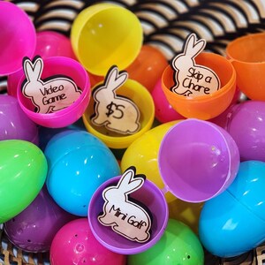 May include: A collection of colorful plastic Easter eggs with wooden bunny cutouts inside. Each bunny has a different message written on it, including "Video Game", "$5", "Skip a Chore", and "Mini Golf".