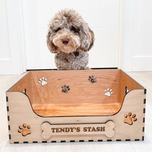 May include: A wooden dog bed with paw prints and the text "Tendy's Stash" on the side. A brown and white dog is looking into the bed.