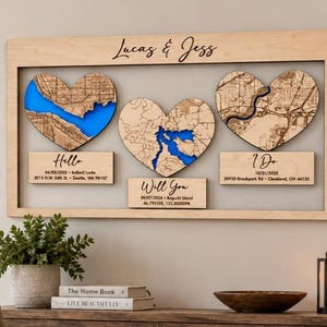 May include: A rectangular wooden wall art piece featuring the text Lucas & Jess at the top. It contains three heart-shaped cutouts with blue water map details and three rectangular plaques below them labeled Hello, Will You, and I Do with dates and addresses.