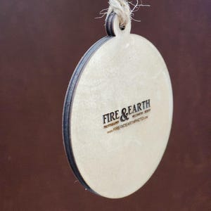 May include: A round wooden ornament with a brown string for hanging. The ornament has a white background with the text "FIRE & EARTH PHOTOGRAPHY" and "www.FIREANDEARTHPHOTO.com" printed in black.
