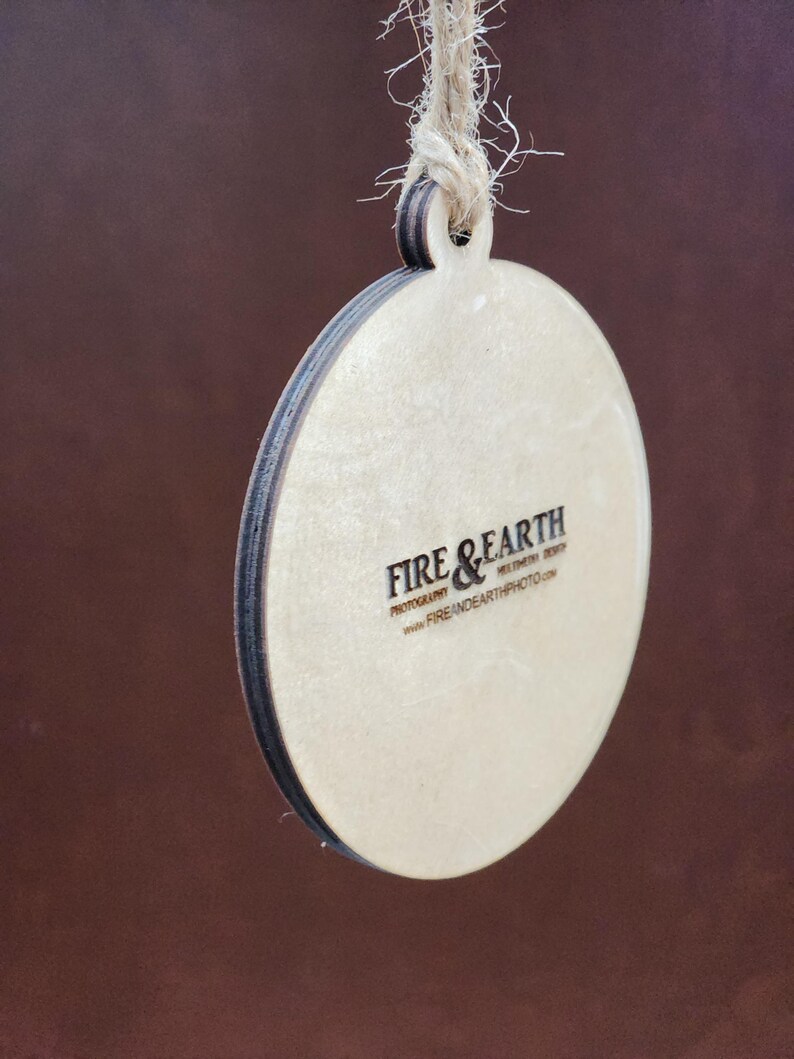 May include: A round wooden ornament with a brown string for hanging. The ornament has a white background with the text "FIRE & EARTH PHOTOGRAPHY" and "www.FIREANDEARTHPHOTO.com" printed in black.