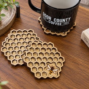 May include: A set of wooden drink coasters shaped like honeycombs with a light brown finish. Each coaster features a hexagonal grid pattern with a small, detailed illustration of a bee with yellow and black stripes on the bottom right corner.