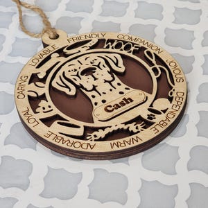 May include: Wooden dog ornament with a brown and white spotted dog silhouette. The ornament has the word "WOOF" and the name "Cash" on it. The ornament is surrounded by words describing dog traits, such as "loyal", "caring", "friendly", and "adorable".