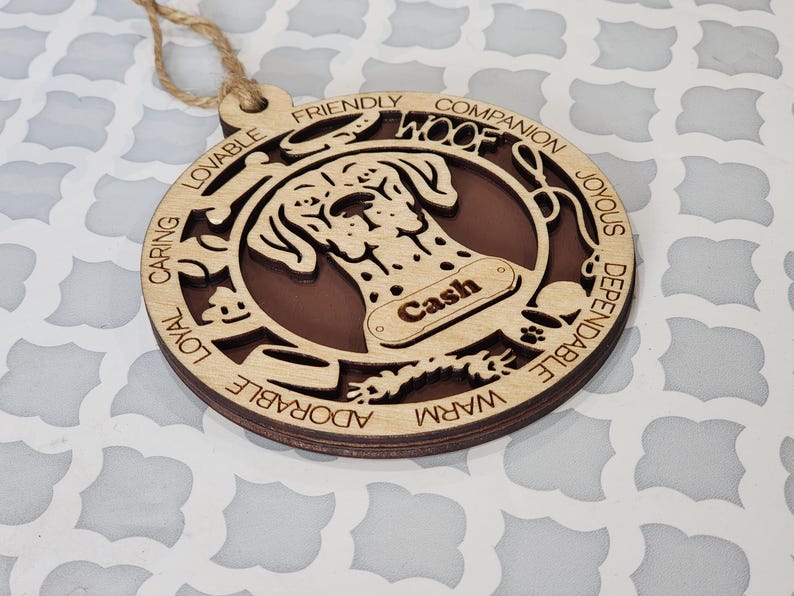 May include: Wooden dog ornament with a brown and white spotted dog silhouette. The ornament has the word "WOOF" and the name "Cash" on it. The ornament is surrounded by words describing dog traits, such as "loyal", "caring", "friendly", and "adorable".