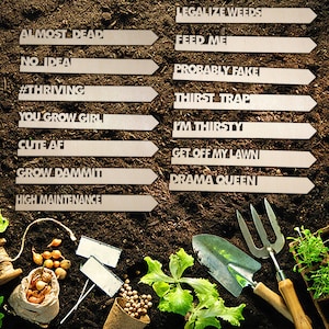 May include: A collection of garden plant markers with humorous and sarcastic phrases like "Almost Dead", "No Idea", "#Thriving", "You Grow Girl", "Cute AFI", "Grow Dammit", "High Maintenance", "Legalize Weeds", "Feed Me", "Probably Fake", "Thirst Trap", "I'm Thirsty", "Get Off My Lawn", "Drama Queen" and "And Many More!" The markers are made of wood and have a white background with black lettering. The markers are arranged in a row on a bed of brown soil. There are also gardening tools, plants, and other gardening supplies in the image.