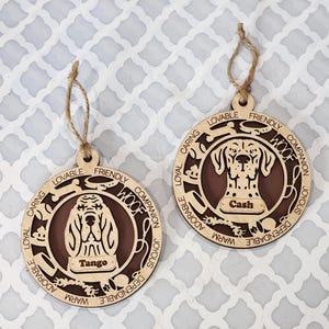 May include: Two wooden dog ornaments with brown painted centers. The ornaments feature a dog's head with the words "Woof", "Loyal", "Caring", "Friendly", "Companion", "Joyous", "Adorable", "Dependable", and "Warm" surrounding the dog's head. The ornaments have the names "Tango" and "Cash" on them.