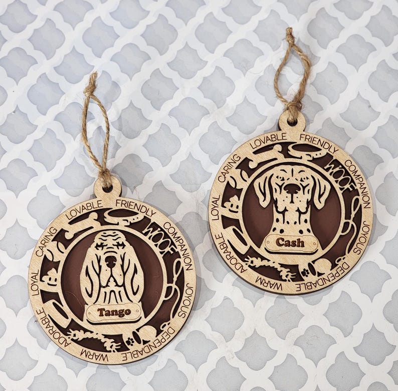 May include: Two wooden dog ornaments with brown painted centers. The ornaments feature a dog's head with the words "Woof", "Loyal", "Caring", "Friendly", "Companion", "Joyous", "Adorable", "Dependable", and "Warm" surrounding the dog's head. The ornaments have the names "Tango" and "Cash" on them.
