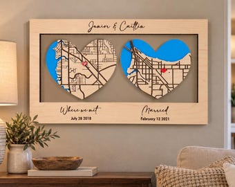 May include: A rectangular wooden wall sign featuring the names Junior & Caitlin at the top. Two heart-shaped cutouts display blue and tan street maps with small red heart markers. Below, the text reads Where we met July 28 2018 and Married February 12 2021.