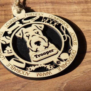 May include: Wooden Christmas ornament featuring a silhouette of a dog with the name "Trooper" on a tag. The ornament is decorated with the words "Loyal, Caring, Lovable, Friendly, Companion, Joyous, Dependable, Warm, Adorable, Woof".