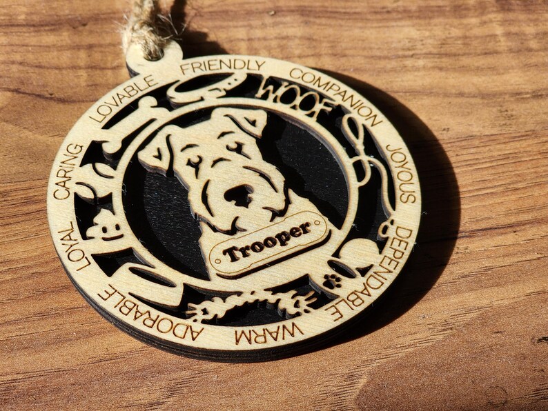 May include: Wooden Christmas ornament featuring a silhouette of a dog with the name "Trooper" on a tag. The ornament is decorated with the words "Loyal, Caring, Lovable, Friendly, Companion, Joyous, Dependable, Warm, Adorable, Woof".